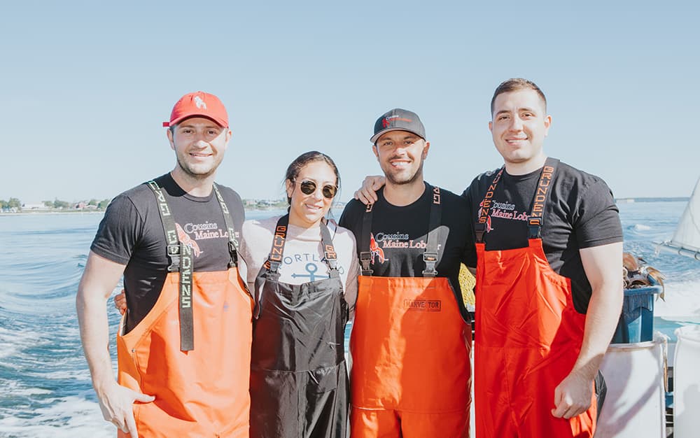 A photo of our Trenton Philly owners on a lobster boat in Maine, in fishing gear, posing with Cousins Maine Lobster founders Jim & Sabin.
